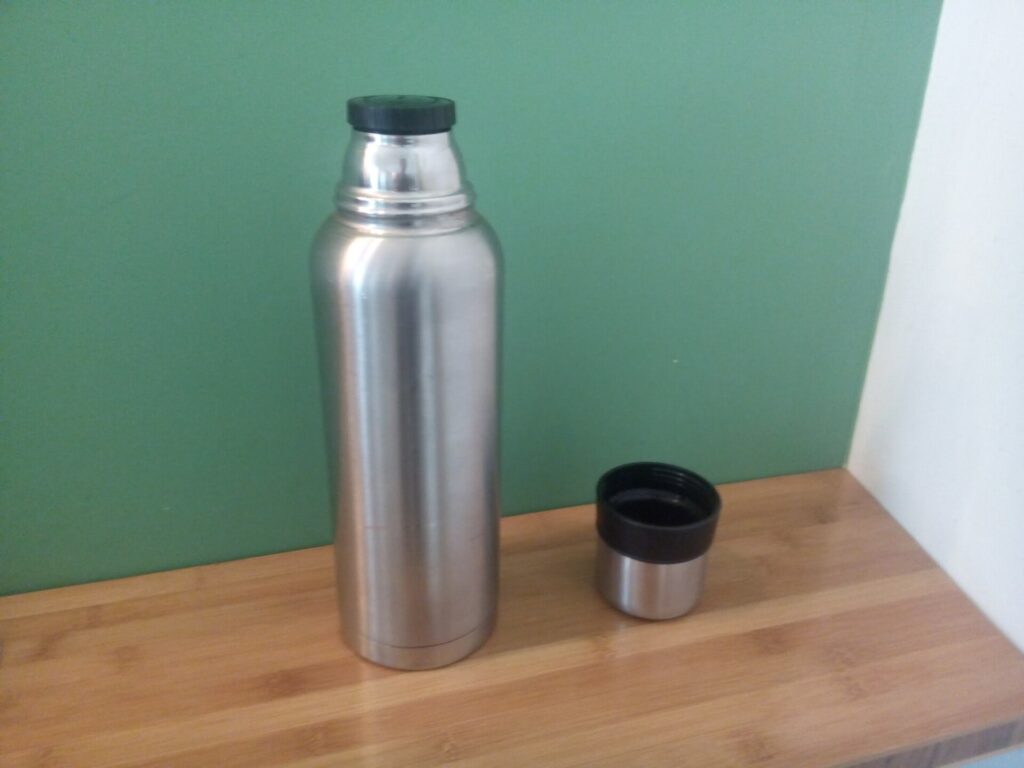 Isolierflasche und Becher mit dampfendem Tee stehen auf einem Baumstumpf im Herbstlaub