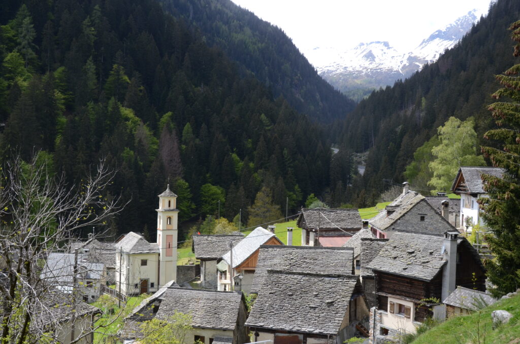 Terrassendorf aus Steinhäusern hoch über einem Alpental, dahinter bewaldete Hänge im Gegenlicht